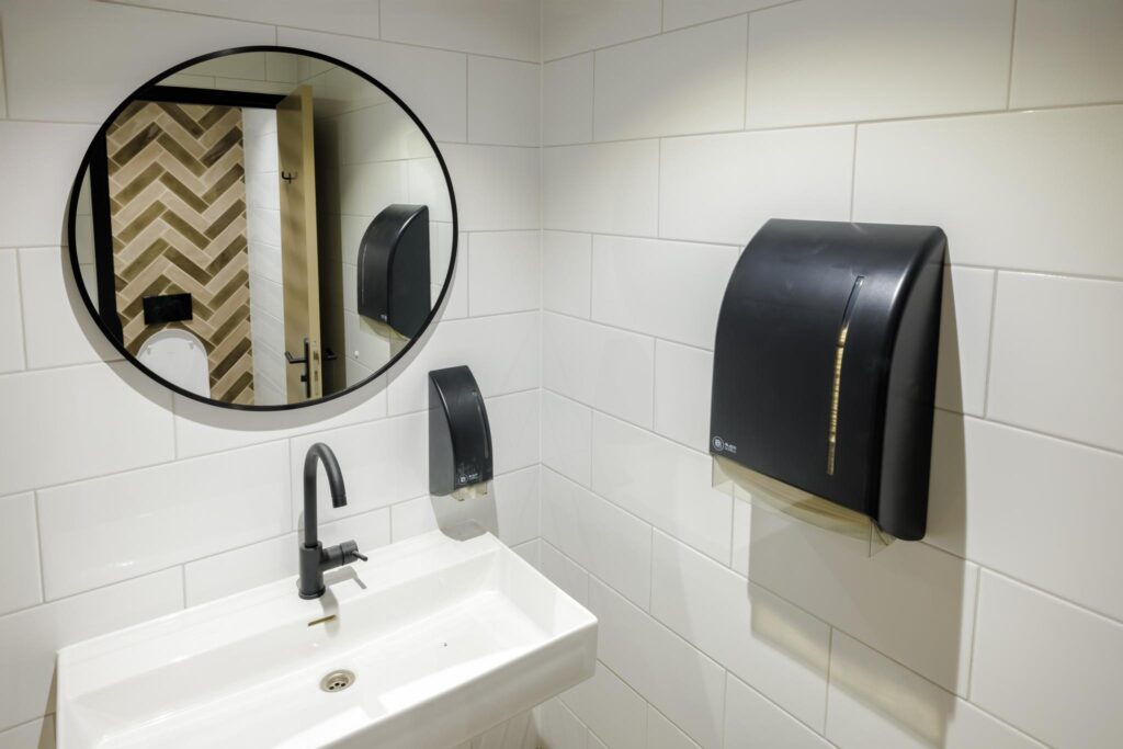 Modern restroom with a white tiled wall, black fixtures, a rectangular sink, and a round mirror reflecting a chevron-patterned wall.